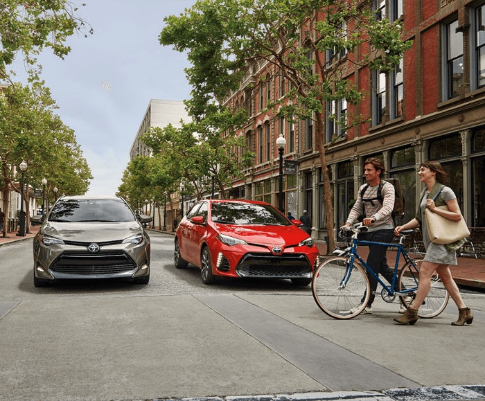 Front-facing view of the 2017 Toyota Corolla LE and XSE in Classic Silver Metallic and Barcelona Red Metallic stopped at a crosswalk in an urban area. 