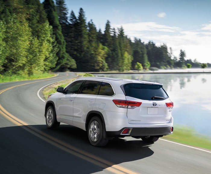 Rear action shot of the 2019 Highlander Hybrid Limited in Blizzard Pearl driving along a forest road.