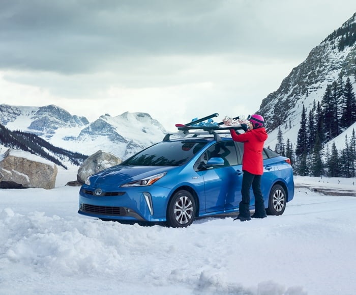 Person removes cargo from Storm Blue Prius roof amid snowy mountain backdrop.