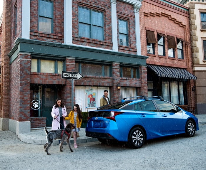 Family and large dog exit cafe to enter 2019 Prius in Storm Blue on city street.