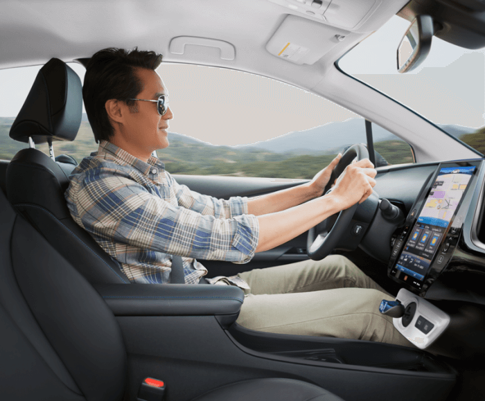 Lateral view of a man smiling and wearing sunglasses while driving the Prius Four Touring. The interior fabric for the seats and front console is black with the ceiling and sun visors in white.  