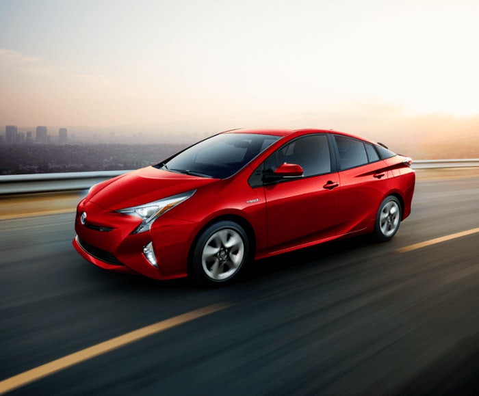 Exterior view of the Prius Four Touring in red being driven with an urban landscape in the background.  
