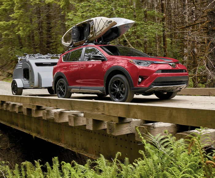 Dual shot of the RAV4 Adventure in Ruby Flare Pearl and the RAV XLE in Magnetic Gray Metallic driving on a forest trail over a bridge. The RAV4 Adventure is carrying a kayak on its roof rack and pulling a trailer. 