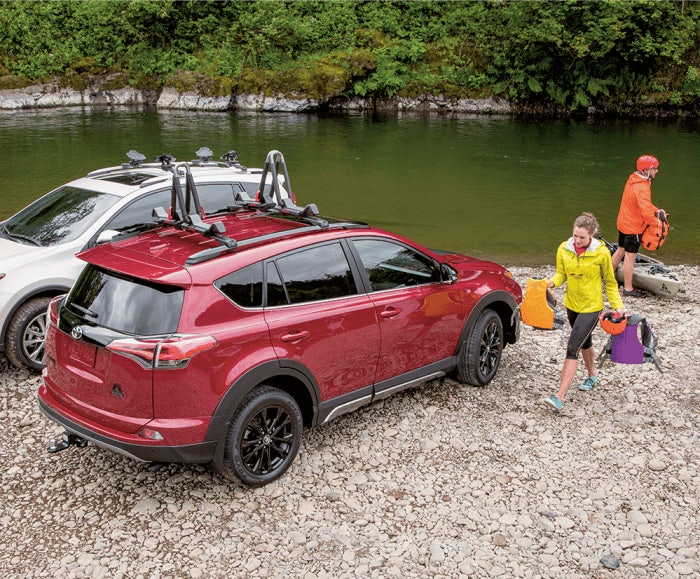 Rear angled view of the RAV4 XLE in Ruby Flare Pearl parked by a river with a woman holding lifejackets by the passenger-side door. 