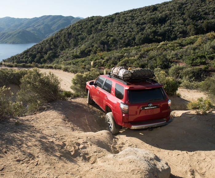 Rear angle of 2019 4Runner in Barcelona Red Metallic driving along rocky, woodsy terrain.