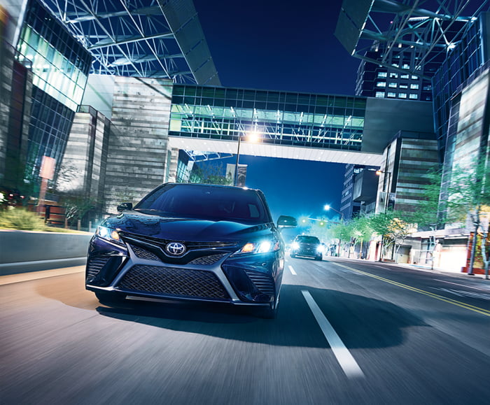 Front exterior shot of two 2019 Camrys in Midnight Black Metallic driving on road at night in front of office builidngs.