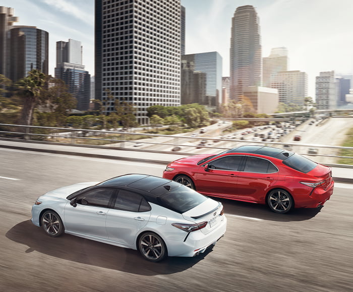 Driver side action profile shot of 2019 Camrys XSE, one in Ruby Flare Pearl and one in Wind Chill Pearl/Midnight Black Metallic Roof.