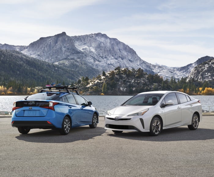 Two 2019 Priuses in Storm Blue and Blizzard Pearl parked beside a lake amid snowy mountains.