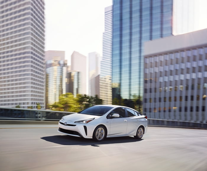 Driver side exterior view of 2019 Blizzard Pearl Prius driving along city street with buildings in background.