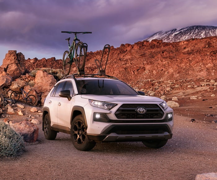 Front view of 2019 RAV4 in Super White with bicycle on roof rack parked in front of mountain.