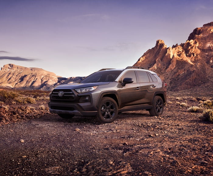 Driver side view of 2019 RAV4 in Magnetic Grey Metallic parked on dirt road in front of mountains.
