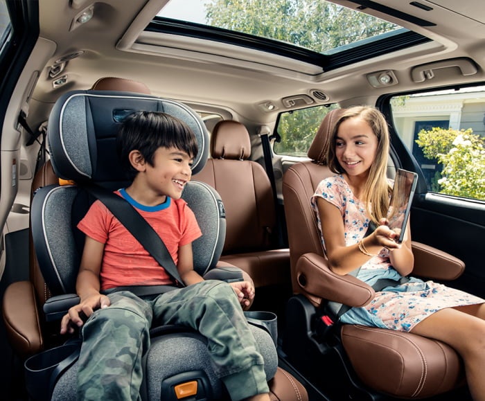 Side angled shot of two children -- one in a carseat in the middle row of 2019 Sienna Limited Premium interior shown in Chestnut with white accent stitching.
