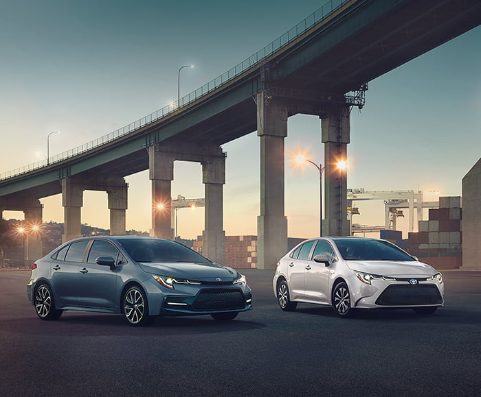 Exterior front 3/4 view of 2020 Corolla XSE in Celestite Gray Metallic and exterior front 3/4 view of 2020 Corolla LE HV in Blizzard Pearl on road underneath freeway.