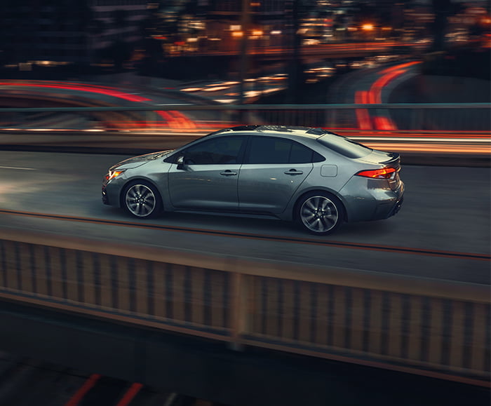 Exterior rear 3/4 driver side view of 2020 Corolla XSE in Celestite Gray Metallic on road with city lights in background.