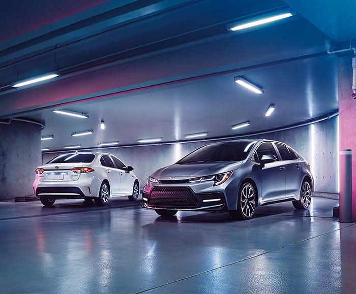 Exterior rear passenger view of 2020 Corolla LE in Blizzard Pearl and exterior front driver view of 2020 Corolla LE in Celestite Gray Metallic in underground parking garage.